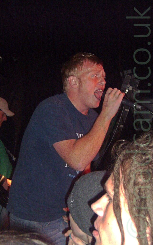 A man with short brown hair, wearing a blue t-shirt with some indistinct text on the front, singing into a microphone held in his right hand. The heads of a couple of people in the crowd can be seen in the foreground, with a man wearing a green t-shirt and a white baseball cap in the background on the left of the frame. Black walls and ceiling fill the rest of the image.