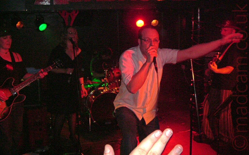 5 people on a stage performing as a rock band. On the left is a woman wearing a black hat and black top with dark trousers, playing an electric guitar. Next is a woman with long blonde hair, wearing red felt reindeer antlers on her head, and a black dress, standing behind a microphone on a stand to do back-up vocals, holding a drink in a bottle in her right hand. Right at the back is a man with long dark hair and wearing a black t-shirt, sitting behind a black drum kit. Right at the front of the stage is a man with short dark hair and eye-glasses, wearing a white shirt and dark trousers, holding a microphone to his mouth with his right hand and his left hand gesticulating to the crowd as he sings On the right is a man in a sparkly stetson, wearing a black t-0shirt and stripey pants, and p[laying a black electric bass guitar. Several fingers belonging to the right hand of someone in the crowd are held aloft in the foreground. Black walls and ceiling fill the rest of the frame, with several coloured lights, mostly red or orange, with one green light, illuminate various parts of the stage, the whole thing having a slight red cast.