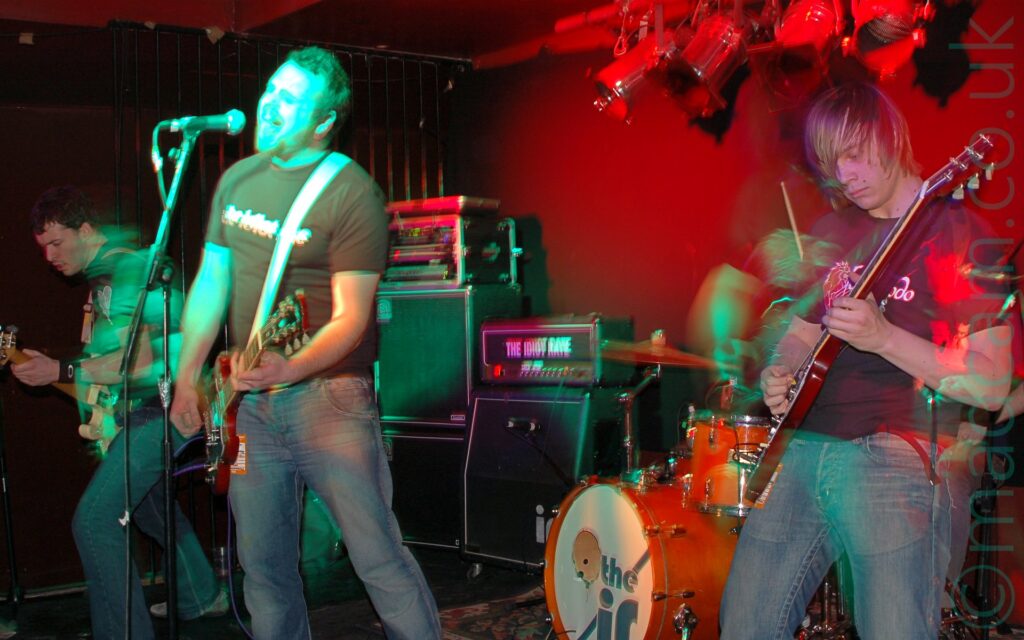 Three people on a stage, performing as part of a band. On the left is a man with short dark hair, wearing a black t-shirt and blue jeans, and playing a white bass guitar while slightly hunched over. In the middle is a man with short dark hair and beard, wearing a dark t-shirt with white text "The Idiot Rate" across the front and blue jeans, playing a red electric guitar while singing into a microphone on a stand in front of him. On the right is a man with chin length dark hair, wearing a black t-shirt with a red and white logo, playing a red electric guitar. I red drumkit can be seen in the background on the right, with a white cover with grey text "The IR" on the front of the bass drum. Stacks of speakers and amplifiers are stacked in front of the red wall in the background on the left, one of which has white "The Idiot Rate" text on the front. A long exposure has combined with their movement and the stage lights to create green and red secondary images and smears of light around the people and equipment.