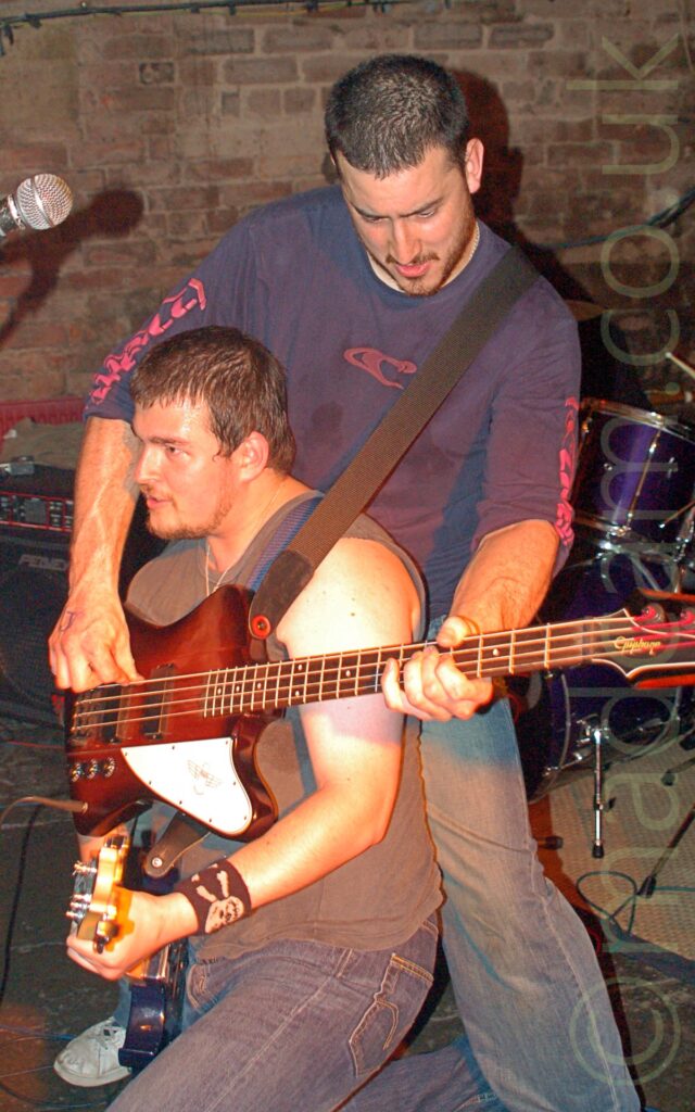 2 members of a band performing on stage, and indulging in some hi-jinks. On the left is a man with short, dark hair and a straggly beard, wearing a grey vest and a blue pair of jeans, crouching down on the floor, holding an electric guitar and facing off to the left. Behind him, on the right, is another man with short dark hair and beard, wearing a blue sweater with red logos on the chest and sides of the arms, and a faded blue pair of jeans. He is holding a red wood and white bass guitar, which is wrapped around the other man. A black and chrome drum kit is in the background on the right, with a brown brick wall filling the rest of the frame.