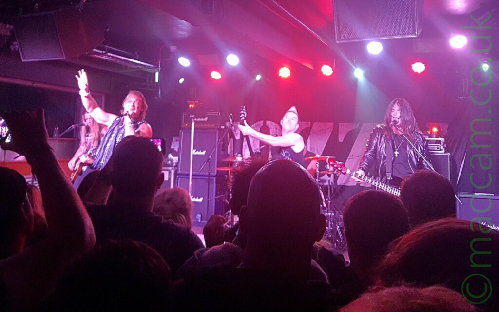 5 members of a band on stage. On the left is a man with long hair, wearing a black t-shirt, and playing an electric guitar. To his right and slightly obscuring him is a man with shoulder-length hair, wearing a sleeveless jacket and blue scarf, with his right arm raised in the air and a microphone held in his left hand, which he is singing in to. In the centre of the image is a man with short blond hair teased up into a single spike, wearing a black sleeveless t-shirt, and playing a electric guitar which is being swung around with reckless abandon. Behind him, almost hidden away, is a man wearing a sleeveless black t-shirt, sitting behind a drum kit. On the right is a man with long, dark hair, wearing a leather jacket over a black t-shirt, and with a large cross on a chain around his neck, playing an inverted bass guitar in a left-handed position. He is singing into a microphone on a stand in front of him. The heads of people in the crowd fill the foreground, with a black backdrop with the white text "Fozzy" stretched between 2 stacks of amplifiers in the background, with bright purple, red, and orange stage lights attached to the black roof above them. The whole scene has a purple cast from these lights.