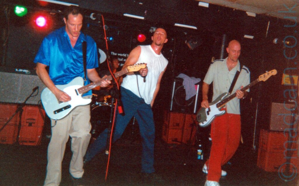 Three men performing on a stage as part of a rock band. On the left is a man with short dark hair, wearing a shiny blue shirt and trey trousers, and playing a silver and white electric guitar. In the middle is a man with a shaven head and short goatee beard, wearing a white tank top and blue trousers, holding a microphone in his right hand. On the right is a man with a shaven head, wearing a grey shirt and red trousers, and playing a silver and grey bass guitar. In the background, speakers are stacked on red crates, with a mi sitting behind a black drum kit visible right at the back of the stage. Black walls and a couple of stage lights fill the rest of the background.