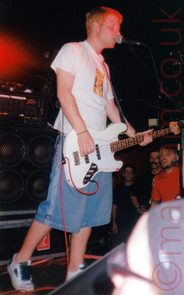 A man, standing facing to the left, with short red hair and sideburns, wearing a white t-shirt with an indistinct yellow logo on the front, and a pair of baggy, knee-length blue jeans and white trainers, playing a white bass guitar while singing into a microphone on a stand in front of him. In the foreground, the head of someone wearing a purple bandana and black eye-glasses can be seen in the bottom right corner. In the background, a group of people can be seen standing at the side of the stage on the right, with stacks of amplifiers on the right. Back walls fill the rest of the background, with a baleful red light in the top left corner.