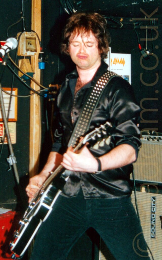 A man with mid-length dark hair, wearing a black silky shirt and black jeans, gurning his way through a song as he plays a black electric guitar, with a heavily studded black strap slung over his left shoulder. Black and yellow amplifiers are stacked on the floor behind him on the right of the frame. Power sockets, wiring runs, and posters adorn the black wall behind him on the left.