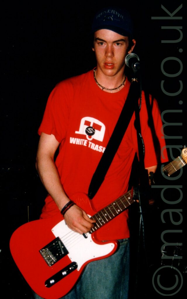 A man with short dark hair, wearing a blue baseball cap, a red t-shirt with a white caravan logo and the words "White trash", and a blue pair of jeans. A black strap slung over his left shoulder supports a red and white guitar he is playing with his left hand. He is standing behind a microphone mounted on a black stand. The space behind him is almost entirely black.