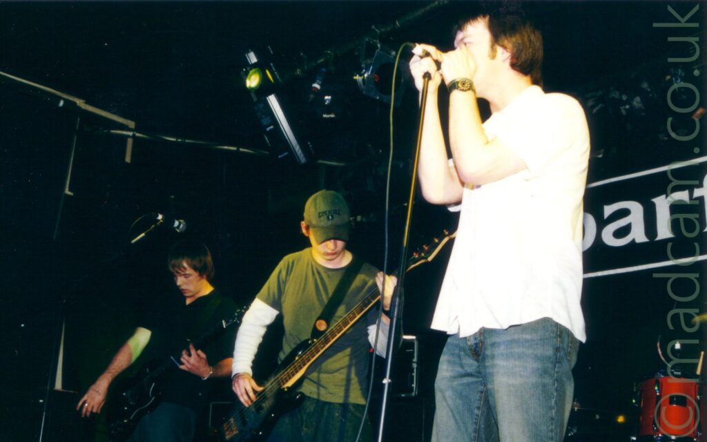 3 members of a band performing on a stage. On the left is a man with mid-length hair, wearing a black shirt and dark blue jeans, playing a black electric guitar, looking down towards his instrument. In the middle is a man wearing a khaki green hat, a green t-shirt over a white jumper, and a blue pair of jeans, playing a black bass guitar. On the right is a man with short brown hair, wearing a white shirt and a blue pair of jeans, holding on to a mic stand as if his life depends on it. Black walls and ceiling fill the rest of the frame, with large white letters "AR" on the rear wall, part of the word "Barfly" on the wall on the right of the frame.
