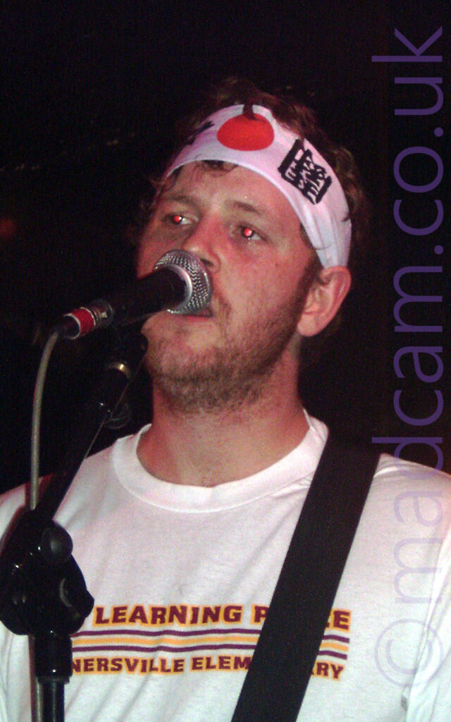 Close-up of the head and shoulders of a man with short, curly hair and short stubble, wearing a wide white headband that appears to have Japanese text on the front, and a white t-shirt with red and yellow text on the front. He is singing into a microphone on a stand, with a black guitar strap over his left shoulder. Black walls and ceiling fill the background.