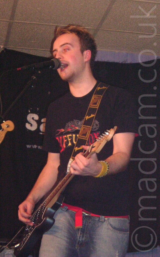 A man with short dark hair, and wearing a black t-shirt with a red and yellow logo across the chest, and a blue pair of jeans with a red belt. He is playing a black electric guitar attached to a black and brown guitar strap with repeated Fender@ logos that goes over his left shoulder, and singing into a microphone on a stand in front of him. Black walls and a white ceiling fill the background.