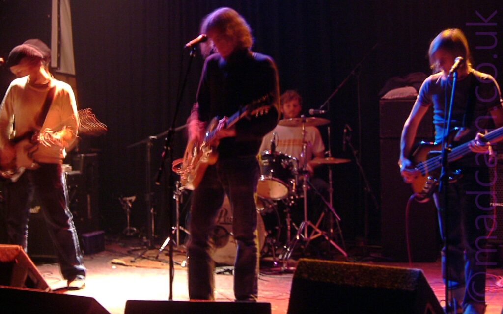 Four members of a band performing on a dark, atmospherically lit stage. On the left is a man wearing a black knitted hat, a white jumper, and dark trousers, playing a black and white electric guitar. In the middle is a man with long, wavy blond hair, wearing a black jumper and dark trousers, playing a brown electric guitar. Behind him is a wan with short dark hair, wearing a white t-shirt and sitting behind a black drum kit. On the right is a man with long, straggly blond hair, wearing a dark t-shirt and blue jeans, and playing a black and brown electric bass guitar. Black curtains fill the background, behind black and grey amplifiers. A long exposure has been used, with movement of heads, arms, and instruments being visible.