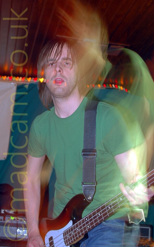 A man with long brown hair, wearing a green t-shirt and a blue pair of jeans, with a black guitar strap slung over his left shoulder attached to the brown bass guitar he is playing. A camera flash has frozen him in place, while a long exposure, a snap zoom, and his movement create secondary images around him. The background has a green and white cloth cover on the wall, with a brown wooden ceiling.