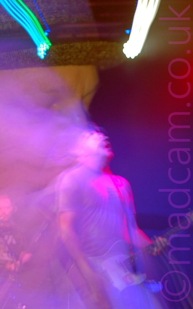 A man on a stage, wearing a grey trilby, a grey t-shirt, and a blue pair of jeans, playing a grey and white guitar with his head thrown back. A long exposure combined with a snap zoom to produce a second image, a close-up of his face on the left of the frame, with streaks of green and white at the top. The whole image has a purple cast, with hints of red.