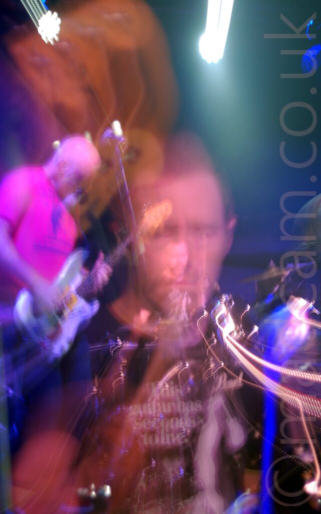 Two men performing on a stage as part of a rock band. The focal point of the image is a close-up of a man with short dark hair and wearing a black t-shirt with white text "This guitar has seconds to live" sitting behind a black drum kit with his eyes closed. A long exposure and a snap zoom out has resulted in a smaller version of his face in the centre of the frame, surrounded by his drum kit, with some streaks of light joining the 2 images. On the left of the frame is a man with a shaven head, and wearing a bright pink t-shirt with an indistinct blue logo, playing a silver and white bass guitar, part of the zoomed-out shot. The head of his bass is visible in the top left corner, coming in to the frame just as the camera zooms out. Blue and yellow smears of light are scattered throughout the frame, with black walls filling the background.