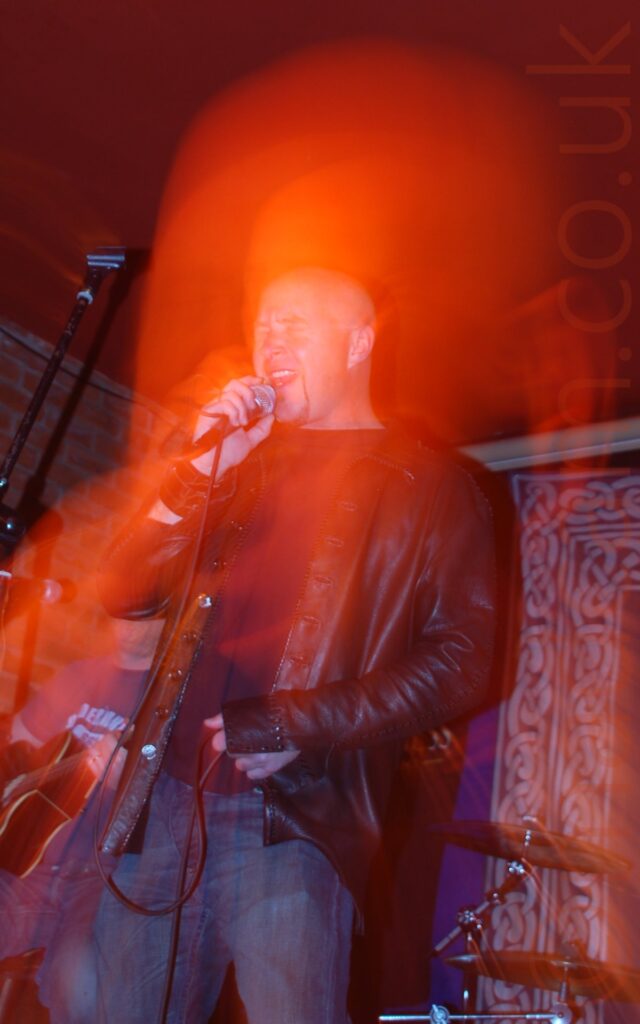 A man with a shaven head and close-cropped beard around his chin, wearing a black leather jacket over a black shirt and blue jeans, as he sings into a microphone held in his right hand, it's lead wrapped around his right wrist. A camera flash has frozen him in place, while a long exposure and snap zoom has created a red halo around his head and upper body, which closer examination shows is actually a close-up of his head. A blue quilt with a border of white Celtic knotwork is in the bottom right corner in the background, with brick walls and red ceiling filling the rest of the frame.