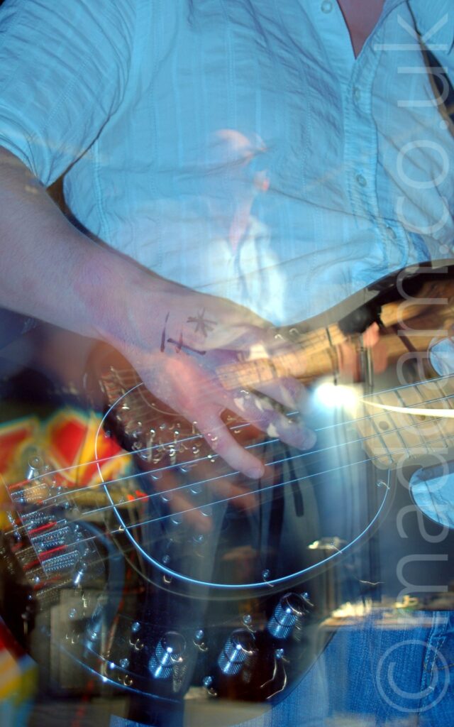 A close-up of a shiny black bass guitar being held by a man wearing a white shirt and blue jeans. A flash has frozen him in place, while a long exposure and snap zoom out conveys movement, with a secondary, smaller image of him holding his bass in the middle of the frame, streaks of lights joining the two images.