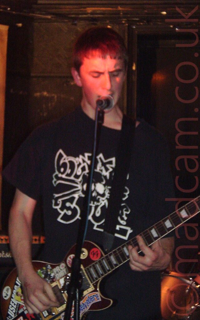 A man on a stage, performing as part of a band. He has short dark hair, and is wearing a black t-shirt with a logo of a skull and cross-bones outlined in white, with several letters scattered around. He is playing a brown electric guitar that has been covered in stickers with other bands logos, and singing into a microphone on a stand. Black walls fill the background.