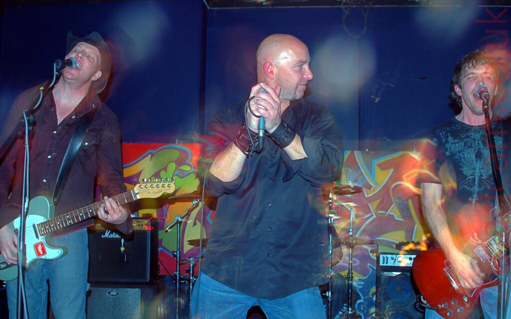 Three men lined up side by side on a stage, performing as part of a band. On the left is a man wearing a black Stetson, a mauve shirt, and blue jeans, playing a grey and white electric guitar as he sings into a microphone on a stand. In the middle is a man with a shaved head, wearing a black shirt with the sleeves partly rolled up, and blue jeans, holding a microphone in his right hand with the cord wrapped around his right wrist while he looks off to towards the man on the right. On the right of the frame is a man with short, slightly wavy dark hair, wearing a black and grey t-shirt and blue jeans, playing a red electric guitar and singing into a mic on a stand. In the background, several amps are standing on the floor, and a drum kit is visible in the middle. A wide but not very tall poster stretches across the frame on the purple walls, with bright, almost psychedelic coloured graffiti.