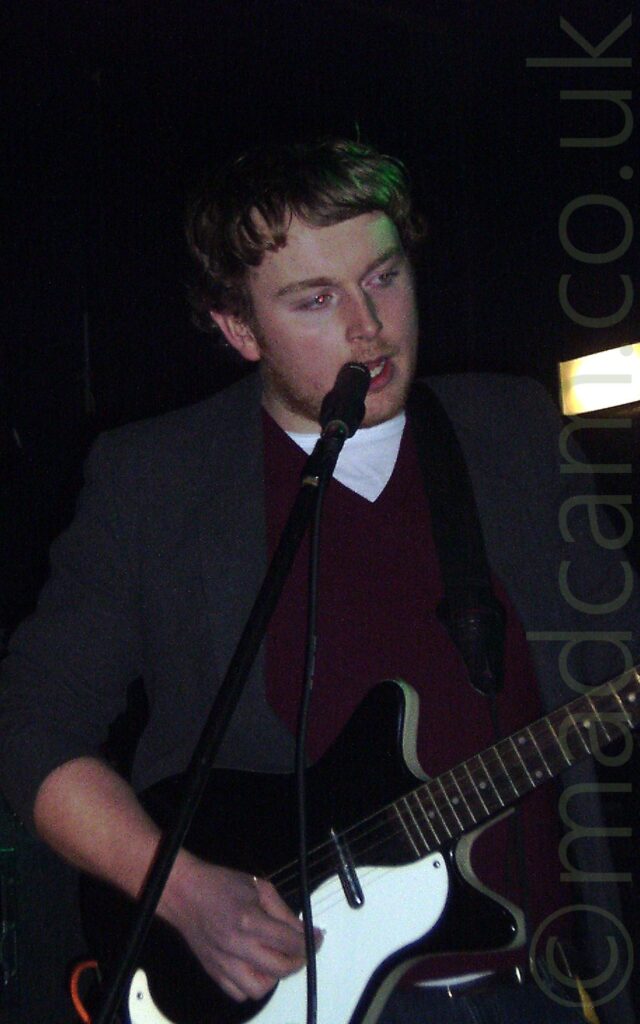 A man with dark hair and a bit of stubble, wearing a purple jumper over a white t-shirt, with a grey suit jacket over the top, playing a black and white electric guitar while singing into a black microphone on a stand. A square white light is shining over his left shoulder, with black walls and ceiling filling the rest of the frame.