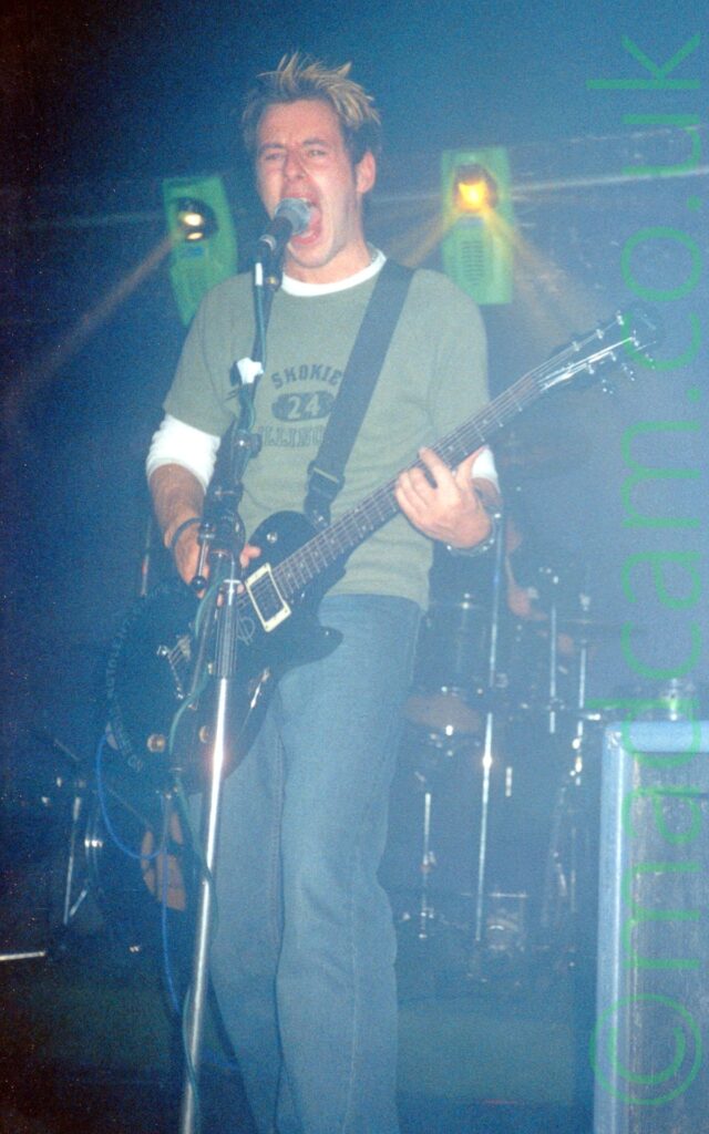 A man with slightly spiky blond hair with dark roots, wearing a green t-shirt over a long-sleeved white t-shirt and blue jeans, playing a black electric guitar attached to a black strap over his left shoulder., while singing (possibly shouting) into a microphone on a stand in front of him. A black drumkit can be seen in the background behind him. A pair of motorised lights in green housings are mounted on the black wall in the background.