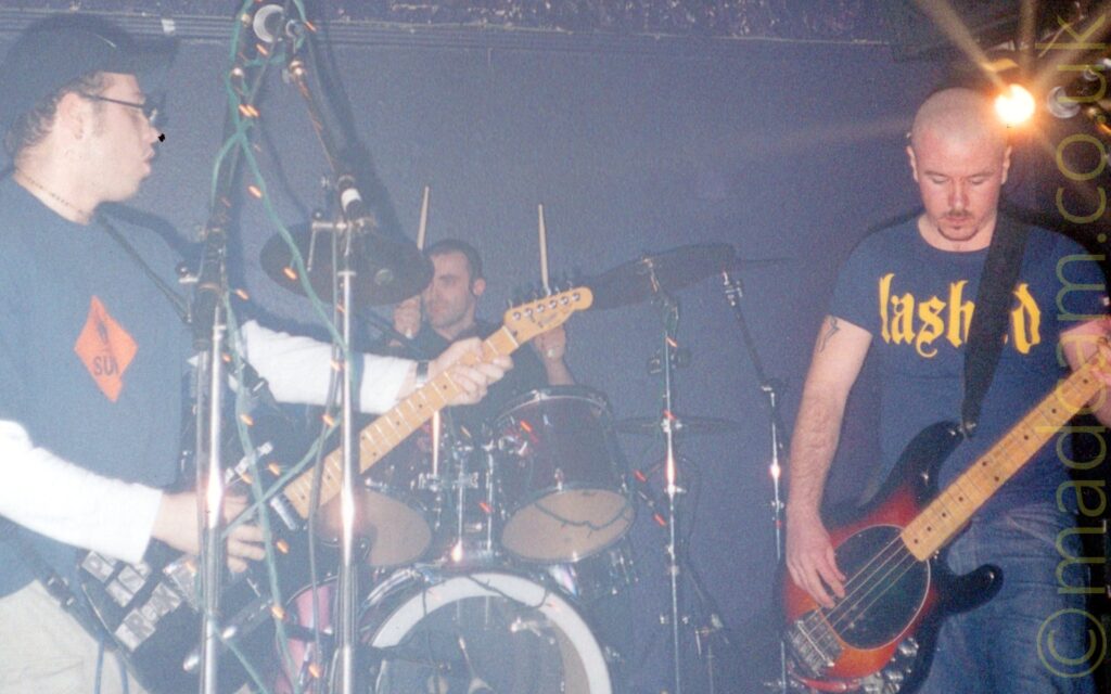 2 men performing on a stage as a band. On the left is a man with a dark blue baseball cap on his head, with a dark blue t-shirt with an orange hazard label over a white t-shirt, and light grey trousers, playing a black electric guitar. In the middle is a man with close-cropped dark hair, sitting behind a black drum kit, with his hands raised in the air holding his sticks aloft. On the right is a man with a shaven head and a short goatee bird, wearing a dark blue t-shirt with the word "lashed" on the front in a yellow, vaguely Gothic font, and a blue pair of jeans, playing a red sunburst and black electric bass guitar. Black walls fill the background, with a bright light shining in the top right corner.