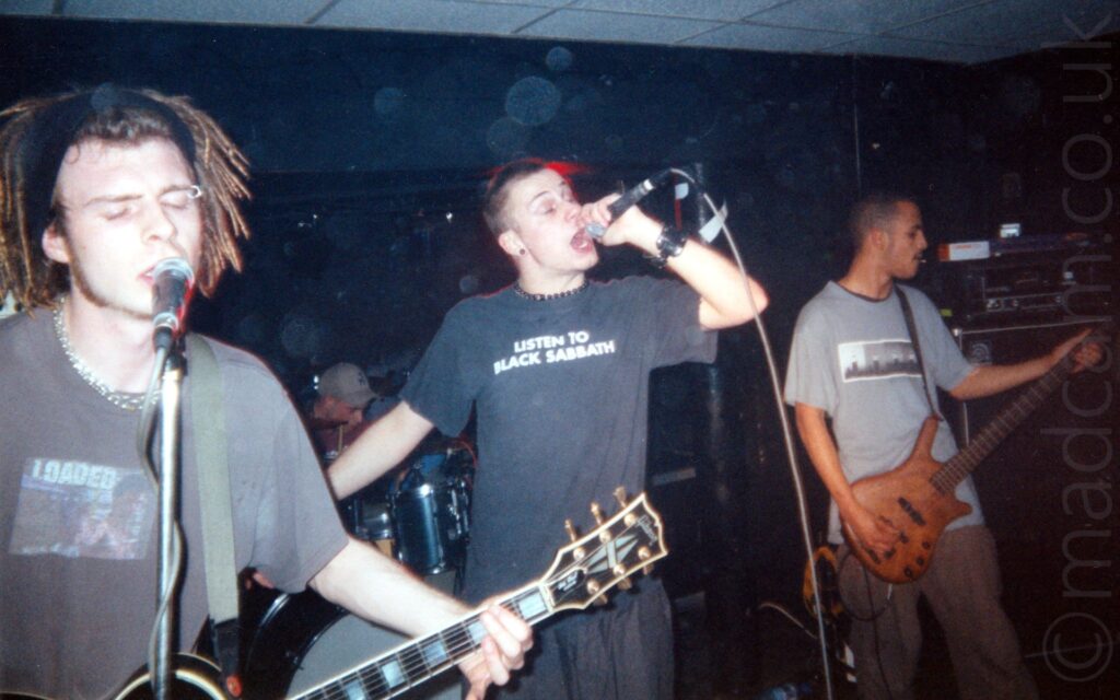 4 men performing on a stage as part of a band. On the left is a man with his hair in dreadlocks, tied back with a black band, wearing a grey t-shirt, and playing a black electric guitar while singing into a microphone on a stand, with his eyes closed. At the back, a man wearing a grey baseball cap is sitting behind a black drum kit. In the middle, a man with short cropped dark hair, wearing a black t-shirt with white text "listen to Black Sabbath", and a black pair of trousers,, holding a microphone in his left hand as he sings, with his eyes closed and mouth wide open. On the right is a man with short cropped dark hair, wearing a grey t-shirt and playing a varnished wooden bass guitar, staring off to his left. A stack of amplifiers is in the background on the right of the frame, with black walls and white ceiling filling the rest of the background.