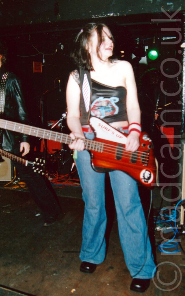 A woman with long dark hair, wearing a black top with a single strap over her right shoulder and an colourful if indistinct logo on the front, and a slightly baggy blue pair of jeans with black boots, playing a red electric bass guitar left-handed, and with a slightly ecstatic grin on her face. Someone wearing a black leather jacket and black jeans can be seen standing on the left of the frame behind her, and amps and other equipment lined up against the black walls in the background. A single green stage light shines out in the top right corner.