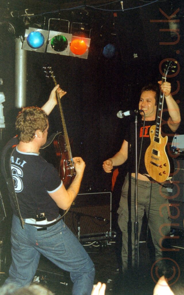 Two men performing on a stage as part of a rock band. On the left is a man, more or less with his back to us, facing to the right of the frame, with short brown hair, wearing a dark blue t-shirt with blue piping on the cuffs of the sleeves and neck, and the blue text "Ali" over a large, white number 6, outlined in light blue, and a blue pair of jeans. He is holding a dark brown guitar aloft in his left hand, while playing with his right hand. On the right is a man with short dark hair, wearing a black t-shirt with a red oblong on the chest, and grey trousers, holding a light brown electric guitar in the air by it's neck in his left hand, as he grins at the other man, with a microphone on a stand in between them. A silvery metal pole holds up the ceiling behind the man on the left, with blue, green, and red stage lights suspended from the ceiling next to it. Black walls fill the background.