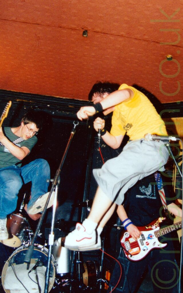 The guitarist and singer from a band jumping exuberantly into the air on stage as they perform. On the right is a young man, with short dark hair and eye glasses, wearing a green t-shirt, blue jeans, and a pair of white trainers, holding a brown electric guitar to one side as he jumps into the air. On the right is another young man, with short dark hair, wearing a yellow t-shirt, grey long shorts, and white trainers, singing into as microphone held in his right hand as he jumps, facing off to the left of the frame. Behind them, on the left, a man wearing a black t-shirt can almost be seen sitting behind a drum kit, with another man wearing a black t-shirt and jeans, and playing a red electric bass guitar, in the background on the right. Black walls and orange ceiling fill the rest of the frame.