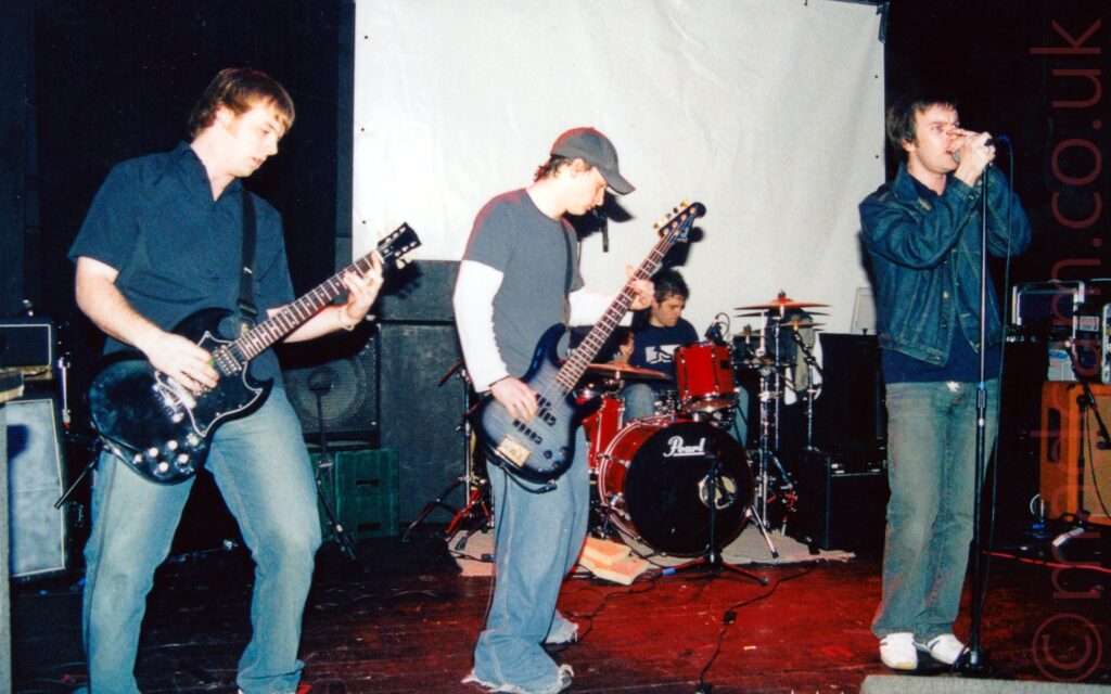 Four men on a stage, performing as part of a rock band. On the left is a man with brown hair, wearing a blue shirt and blue faded jeans, playing a black electric guitar. To the right is a man wearing a grey baseball cap, and a grey t-shirt over a long-sleeved white t-shirt, as well as baggy blue jeans with frayed ends. At the back is a man with short dark hair, wearing a dark blue t-shirt with a white monkey logo and blue jeans, sitting behind a red drum kit, drum sticks held aloft in both hands. On the right is a man with short dark hair, wearing a blue denim jacket over a dark blue polo-shirt and blue jeans, singing into a microphone on a stand that he is clasping in both hands. Stacks of speakers and amplifiers litter the back of the stage, with a white screen mounted on the black wall at the back, just behind the drummer. A red spotlight gives the centre of the stage a red cast.