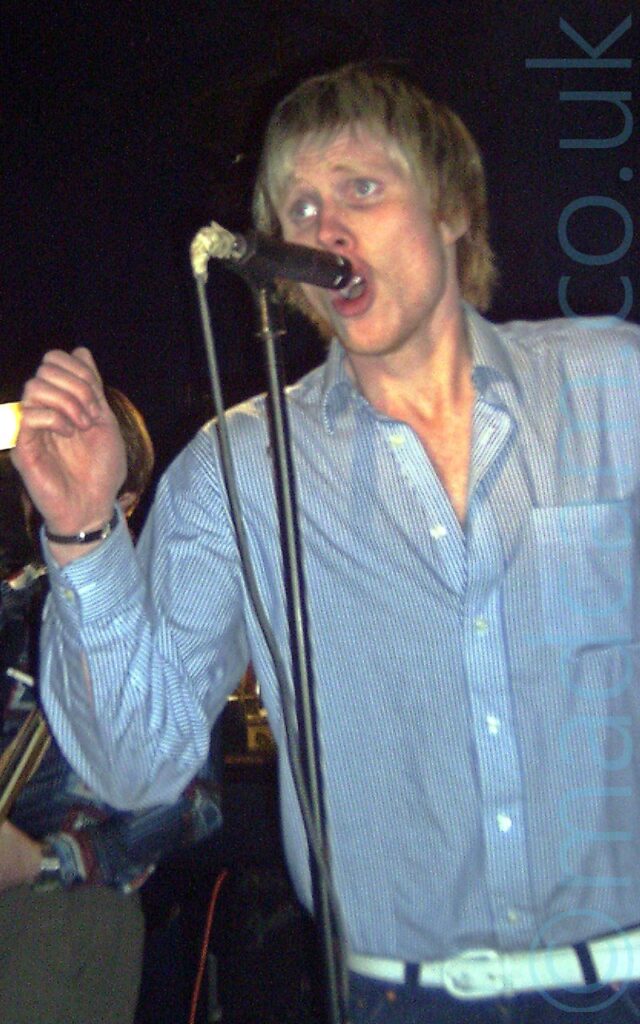 A man with shaggy blond hair, wearing a blue and white pin-stripe long-sleeved shirt, open to the 3rd button, and a blue pair of jeans with a white belt, singing into a microphone on a stand. A man playing an electric guitar is visible in the background on the left of the frame, with black walls and ceiling filling the rest.