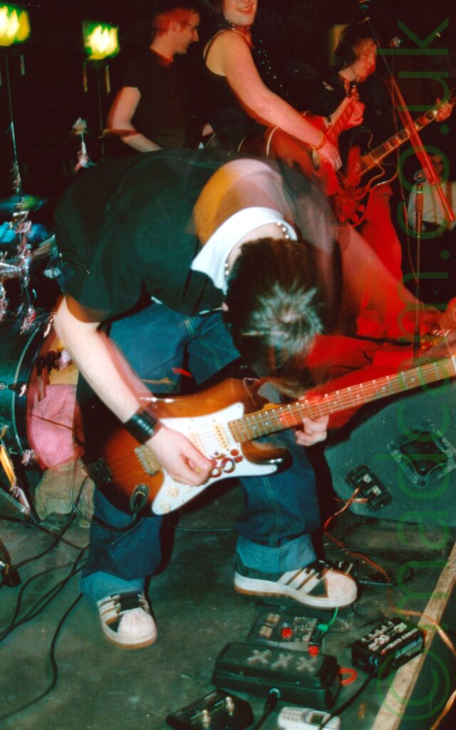 A man performing on a stage as part of a rock band. He has dark hair, and is wearing a green shirt with a white collar, and blue jeans with massive turnups at the bottom. He is hunched over, and playing a brown and white electric guitar, with an array of effects pedals by his feet. A camera flash has frozen him in place, while a long exposure and his movements have created secondary images around his head and arms. A green drum kit is visible on the left of the frame, and a grey wedge speaker on the right. There are 3 people in the background, all members of the band. On the left is a man with short dark hair, and wearing a black t-shirt. In the middle is a woman with her dark hair cut in a bob, and wearing a strappy black top, and looking over at the camera and smiling as she plays an acoustic guitar. On the right is a man wis feather light hair, wearing a black jacket, and playing an electric guitar. The 3 people in the background all have varying degrees of a red cast from the stage lights. A pair of green emergency exit lights shine on the black wall on the left of the frame.