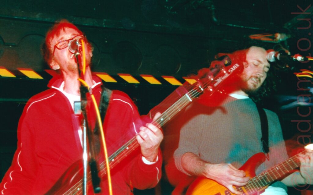 On the right is a man with a veritable mop of long, curly black hair and a close-cropped beard, wearing a grey jumper with the sleeves rolled up almost to the elbows, while playing a varnished wooden electric guitar. Black walls and ceiling fill the background, with yellow and black hazard taps marking the point where the rear of the stage is inset into the wall, creating a low ceiling.
