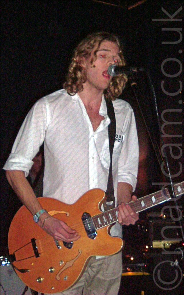 A man with shoulder-length, wavy blond hair, wearing a white shirt and beige trousers, playing and an amber-coloured polished wood electric guitar while singing into a microphone on a stand. Racks of amplifiers and part of a white drum kit can be seen at the bottom of the frame in the background, with black walls and ceiling filling the rest of the frame.
