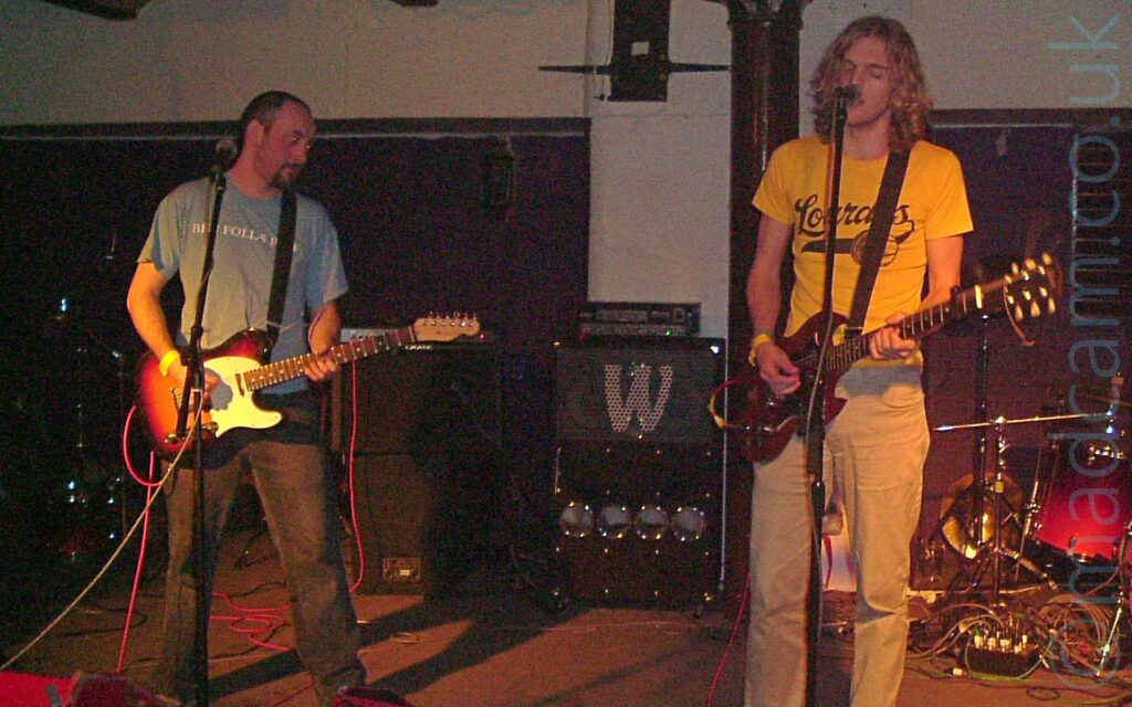 2 men performing on a stage as part of a band. On the left is a man with close-cropped dark hair and goatee beard, wearing a pale blue t-shirt with white "Ben Folds Five" text on the front, and playing a red sunburst and white electric guitar as he stares off to his left. On the right is a man with shoulder-length wavy blond hair, wearing a yellow t-shirt with a black logo, and beige trousers, playing a red electric guitar while singing into a microphone on a stand. A red and chrome drum kit is in the bottom right corner of the background, with an amplifier and a couple of stacked speakers in the middle of the background. The walls in the background are white on top and a deep purple at the bottom.