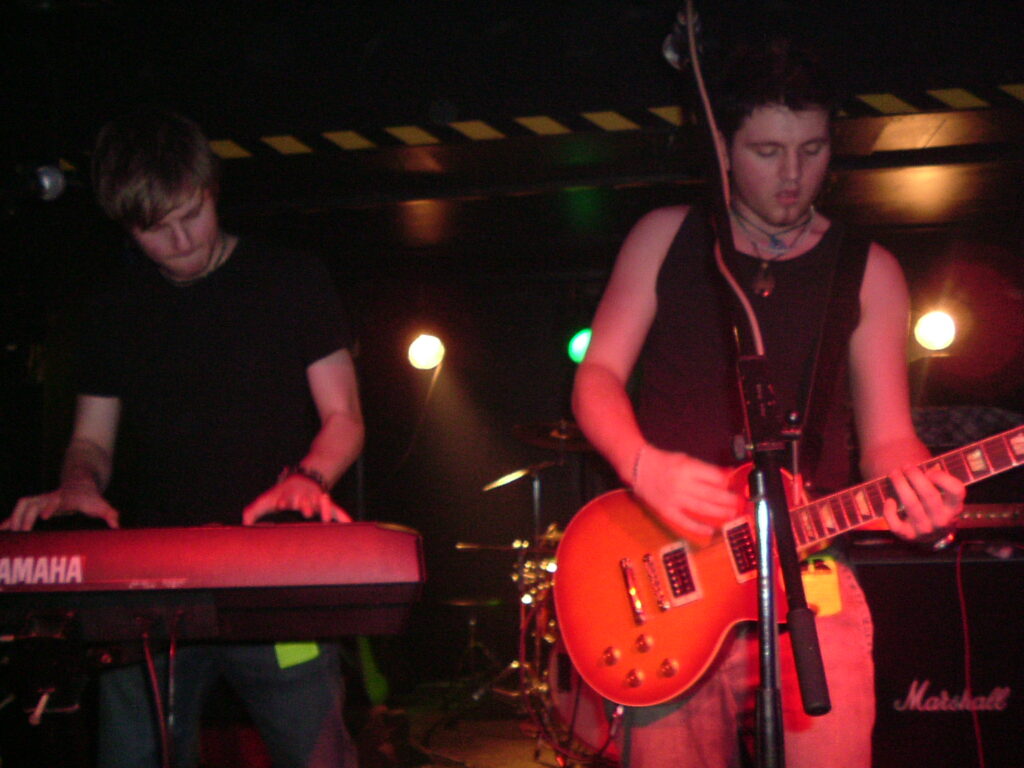 Two men on stage, performing as part of a band. On the left is a man with short dark hair, wearing a black t-shirt, and playing a black keyboard with a white "Yamaha" logo, his hands hovering over the keys. ON the right is a man with short dark hair, wearing a black tank top and jeans, playing an amber, varnished electric guitar. Black walls and ceiling fill the background, with a strip of black and yellow hazard tape marking the low ceiling where the stage is set back into the wall. Bright yellow and green lights shine at the back of the stage.
