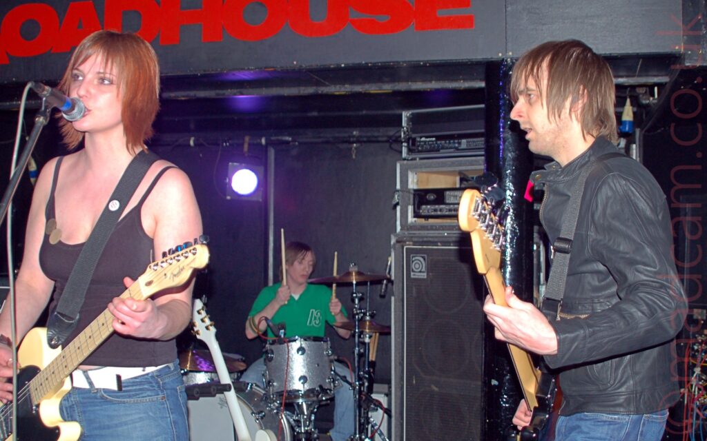 3 people on a stage, performing as a band. On the left is a woman with shoulder-length red hair, wearing a strappy black top and a blue pair of jeans, and playing white electric guitar attached to a black strap over her left shoulder. In the middle is a man sitting behind a silver drum kit at the back of the stage, with brown hair and wearing a green t-shirt, a pair of drum sticks raised in his hands. On the right is a man with shaggy brown hair, wearing a black leather jacket and a pair of blue jeans. He is playing a black bass guitar and facing to the left of the frame. Black walls fill the rest of the frame, with the red word "Roadhouse" on the wall right at the top of the frame.