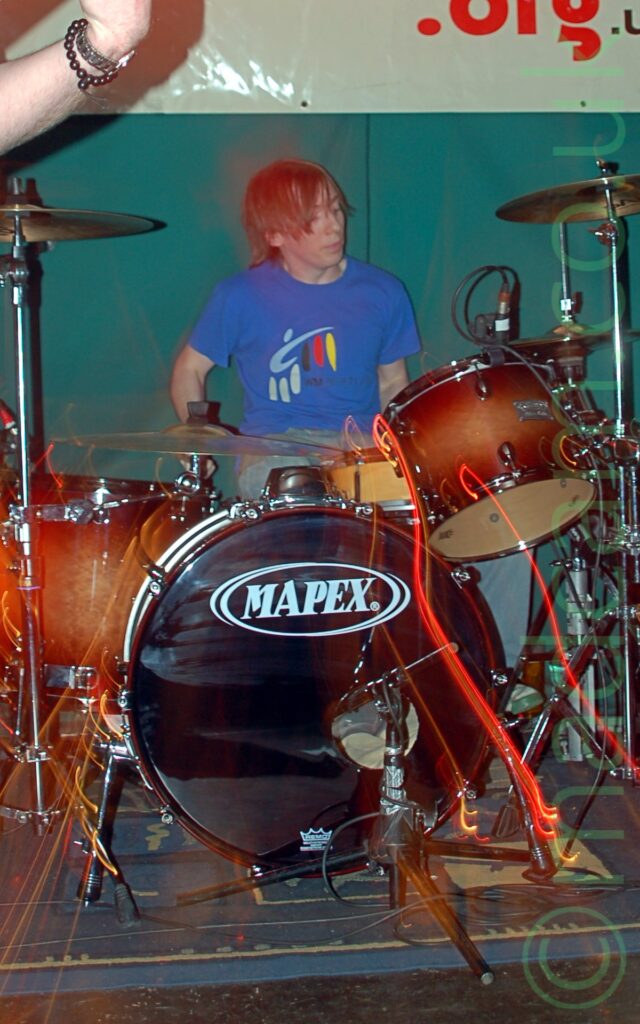 A man with mid-length b4rown hair, wearing a blue t-shirt with a logo made up of light blue, white, black, red, and yellow swirls, sitting behind an amber-coloured drumkit. The fore-arm of a man with a couple of bangles on he wrist is in the top left corner, apparently playing a guitar. Green walls, with a creamy-green plastic banner covering the top, fills the background. Red and orange squiggles of light fill the bottom of the frame, reflecting off the metal parts of the drum kit, suggesting a zoom was used while the photo was being taken.