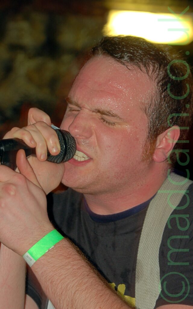 Close-up of the head and shoulders of a man with short dark hair, wearing a dark grey t-shirt, with a light grey guitar strap over his left shoulder. He is clasping a microphone on a stand with both hands as he leans in to sing, and sweating profusely across his forehead. A mottled yellow and black pattern is out of focus on the wall behind him, with a yellow emergency light shining above his head.