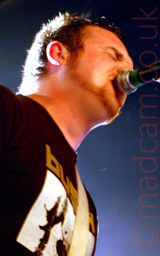 Low shot of a man singing into a microphone on a stand that is almost in his mouth. He has short dark hair and a stubbly beard, and wearing a black t-shirt with a yellow "Bush" logo over an indistinct black and white image. Bright lights in the background backlight his head and body.