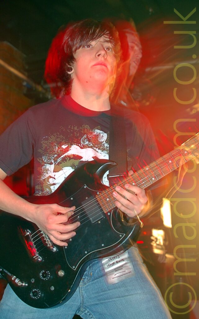 A man with chin length dark hair, wearing a black t-shirt with an extravagant green and white logo filling the chest, and a pair of blue jeans, playing a black electric guitar. A flash from the camera has frozen him in place, but a long exposure and his movement create secondary images, especially around his arms and head. Red stage lights create a red aura around his head and body, while yellow lights give the brown brick walls and black ceiling in the background a yellow cast.