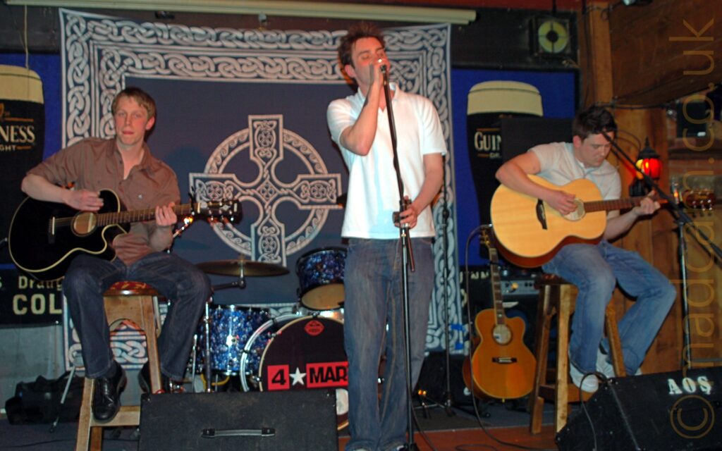 3 men performing on a stage as part of a band. On the left is a man with short blond hair, wearing a brown shirt and a pair of black jeans, sitting on a tall brown stool and playing a black acoustic guitar. In the middle is a man with spiky brown hair and weraing a white polo short and grey jeans, singing into a microphone on a stand, holding the mic in his right hand and the stand in his left. On the right is a man with short dark hair, wearing a white t-shirt and a blue pair of jeans, sitting on a tall wooden stool and playing a brown acoustic guitar. In the background in the middle is a sparkly blue and chrome drum kit, with a large, black "4*Mary" logo in the middle of the bass drum skin, with another brown acoustic guitar off to one side. On the wall is a large, dark blue fabric covering with a large, white Celtic cross in the middles, and bordered with elaborate white scrollwork. Large blue posters featuring over-sized pint glasses filled with a black liquid with a white top, and emblazoned with the word "Guiness" are on either side of the stage.