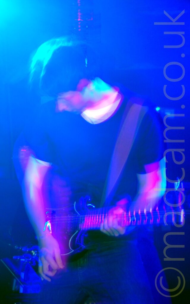 A man on a stage, playing a black electric guitar. He is wearing a black t-shirt over a white t-shirt, and a dark pair of trousers. A long exposure and his movement have combined with the stage light to create secondary images of him, particularly his arms and head, with a blue light in the top left corner giving the whole frame a clue cast.
