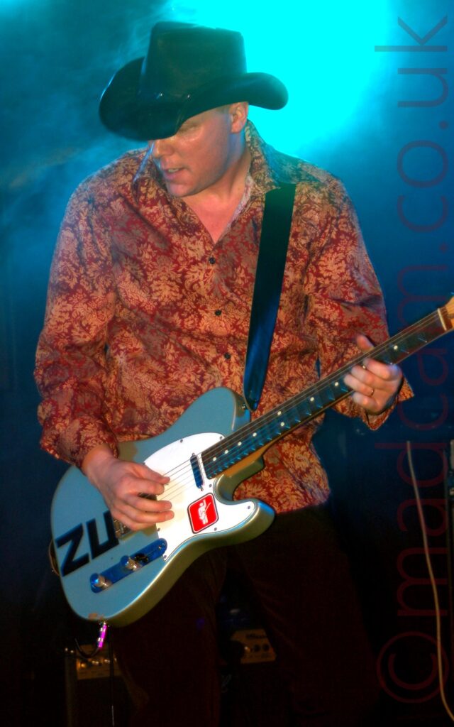 A man on stage wearing a black stetson, a red shirt with an orange flowery print, and a dark-coloured pair of trousers, playing a grey and white electric guitar with the letters "ZU" marked out in tape and a red square logo of a white stick figure holding a guitar with it's left hand raised in the air. A beam of blue light shines down behind him, illuminating the rear of the stage, diffused through a cloud of smoke.
