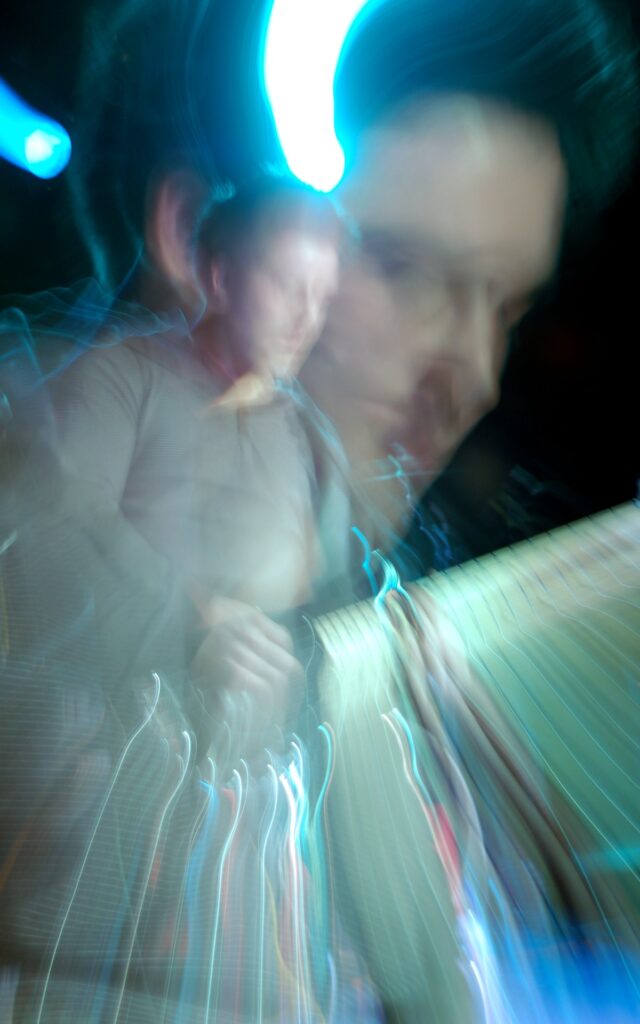 A man with dark, slightly fluffy short hair, wearing a grey polo shirt, and playing a black bass guitar on a stage as part of a band. a long exposure and a snap zoom combines with the stage lights and his movement to produce a secondary, close-up image of his face, and streaks of light, mostly green, coming downwards from his guitar, and a streak of blue light at the top of the frame. The whole image has a slight green and blue tinge.