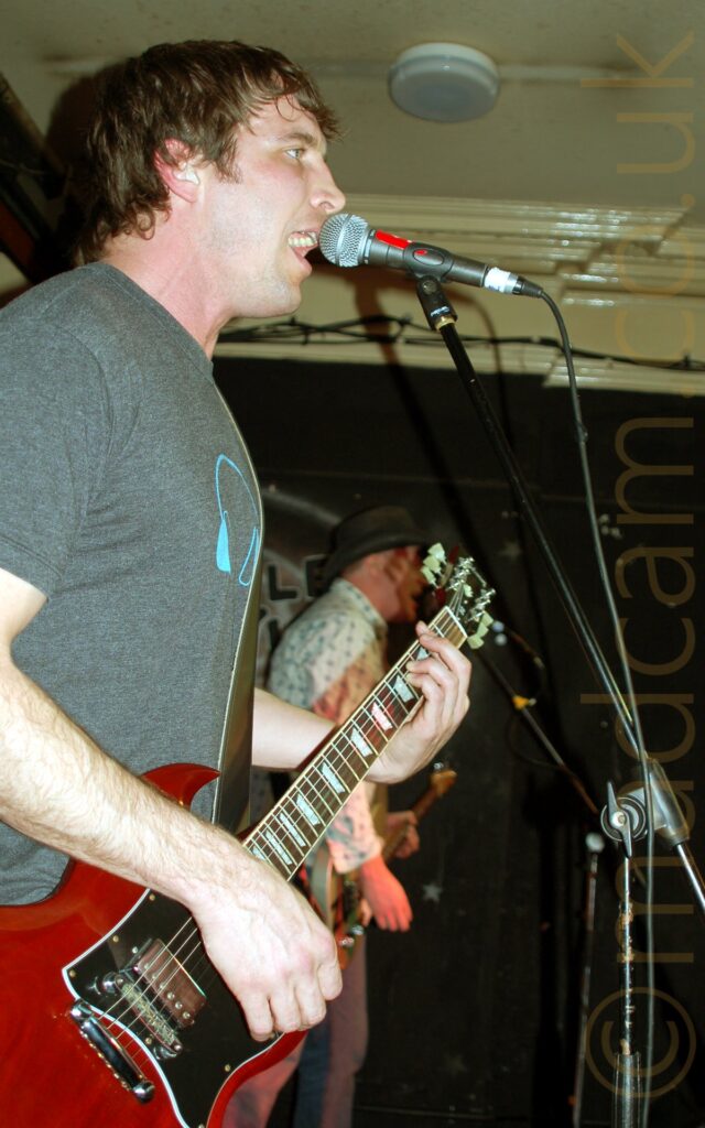 Side view of a man with shaggy brown hair, wearing a grey t-shirt with a blue headphones logo in the middle, facing to the right. He is playing a red electric guitar, and singing in to a microphone on a stand in front of him. In the background is a man wearing a black, wide-brimmer hat, a white shirt with a blue floral pattern, and a pair of grey trousers. He is playing a grey electric guitar, and singing into a microphone on a stand. Black walls and white ceiling fill the rest of the frame.