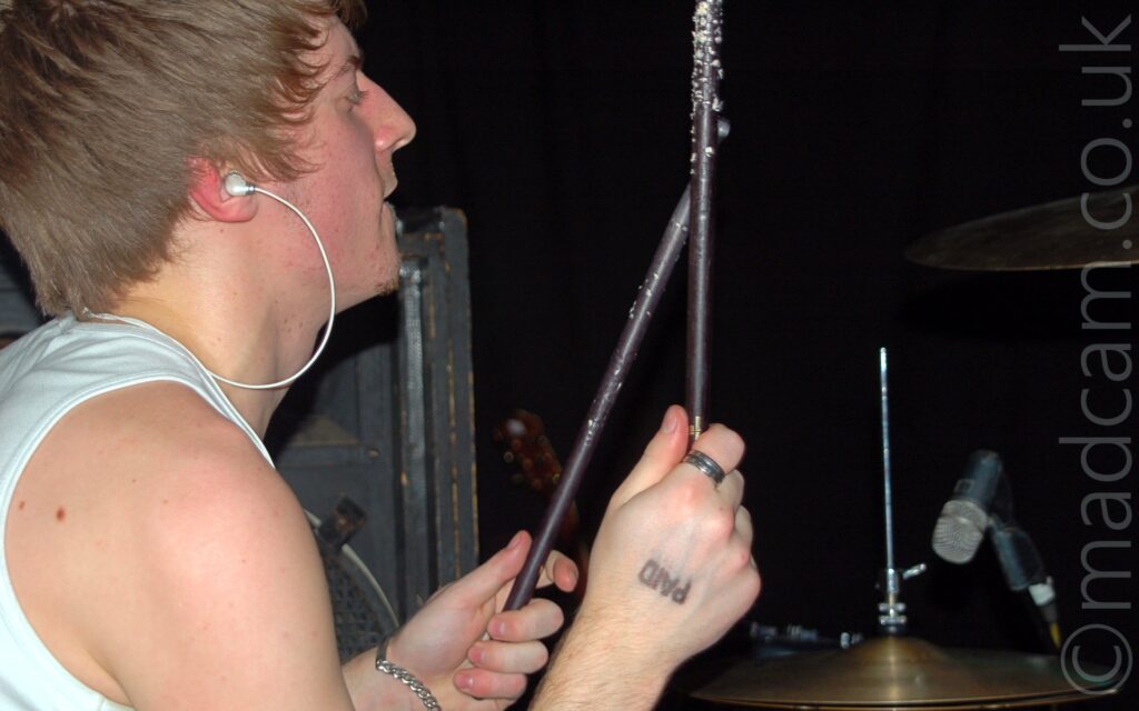 Close-up of the head and shoulders of a man with short brown hair, wearing a white tank top, and with white wired earbuds in his ears, sitting behind a drum kit, with a pair of black drum sticks held in his raised hands. The black word "Paid" has been stamped on the back of his right hand. Black walls fill the background.