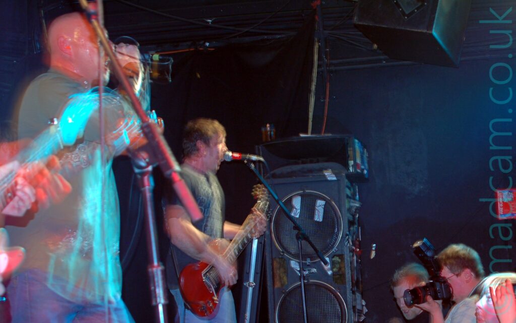 Two men on the left of the frame, performing on a stage as part of a band. On the left is a man with a shaven head, wearing a green long-sleeved shirt and blue jeans, holding a microphone to his mouth with his right hand as he sings, the cord wrapped around his right wrist. Slightly to the right is a man with short dark hair, wearing a grey t-shirt with a large, indistinct white logo on the front, playing a red electric guitar while singing into a microphone on a stand. A tall speaker stack is in the middle of the frame, leaning against the black wall which fills the background. Several people in the crowd can be seen in the bottom right corner, including a man in a white shirt with a large black camera held up to his face, taking photos of something on stage. A camera flash has frozen everyone in place, while a long exposure and their movement has left everyone a bit indistinct, with secondary images and light halos all over the place.