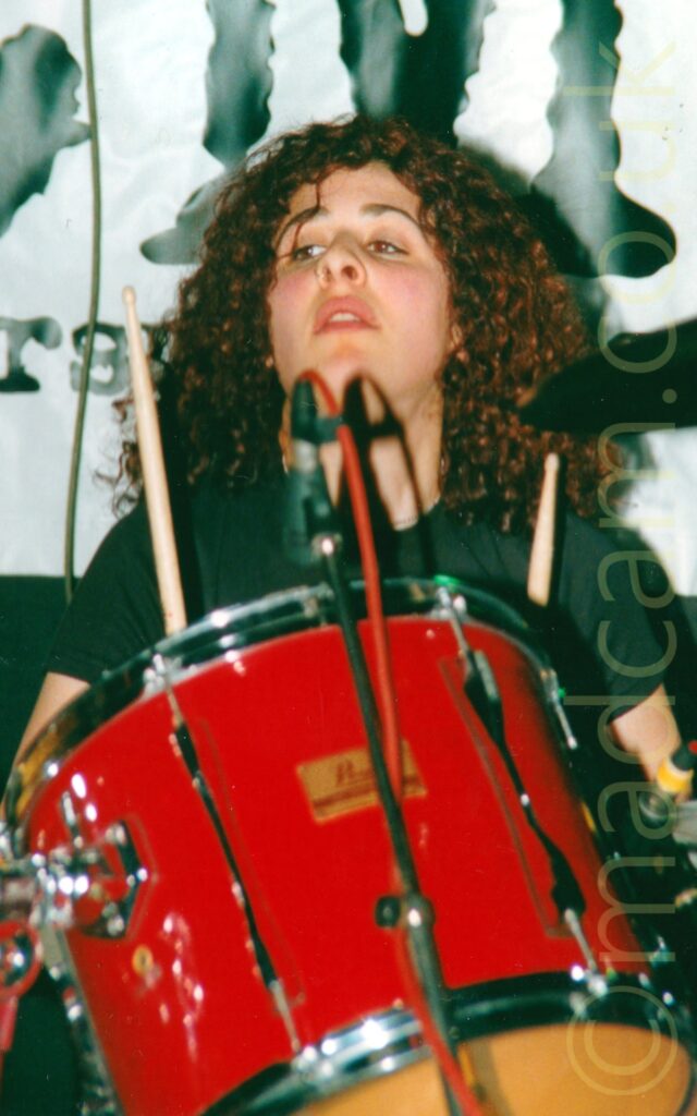 A woman with long, curly brown hair, wearing a black t-shirt, sitting behind a red drumkit. A large white poster with indecipherable black text fills the frame behind her.
