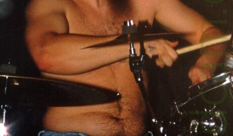 A shirtless man with dark hair and wearing a blue pair of jeans, sitting behind a black and chrome drumkit. His arms are slightly raised as he plays his instrument, slightly leaning back, his head turned slightly to his left with his eyes closed. Black walls fill the background.