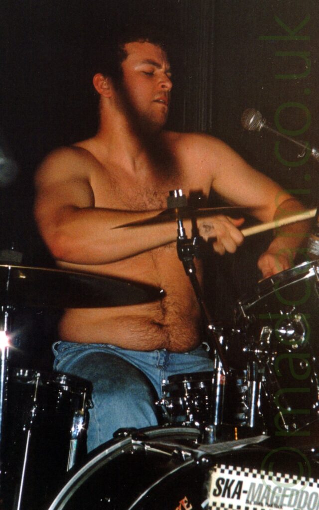 A shirtless man with dark hair and wearing a blue pair of jeans, sitting behind a black and chrome drumkit. His arms are slightly raised as he plays his instrument, slightly leaning back, his head turned slightly to his left with his eyes closed. Black walls fill the background.
