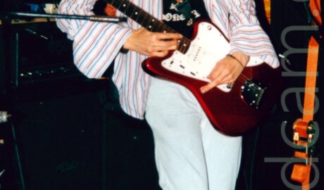 A man with long, wavy black hair, wearing an open white shirt with red and blue pin-stripes and large, open cuffs on the sleeves, over a black t-shirt with a white logo, and flared white jeans, playing a red and white guitar left-handed. He is looking down at his guitar, his face covered with his long hair. A microphone on a stand is in front of him, but he isn't using it. There are a number of black amps scattered around behind him, leaning against the orange and yellow wall, or the black wall at the back of the stage.